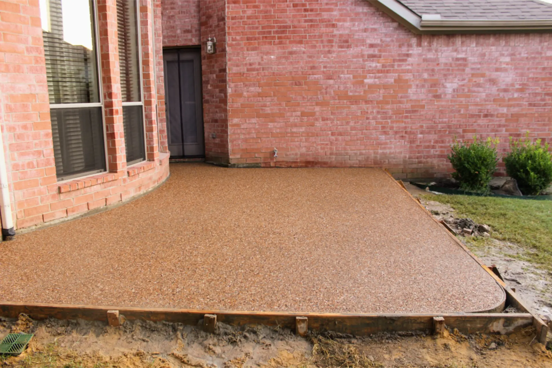 A finished exposed aggregate concrete patio beside a brick home, still edged with timber formwork